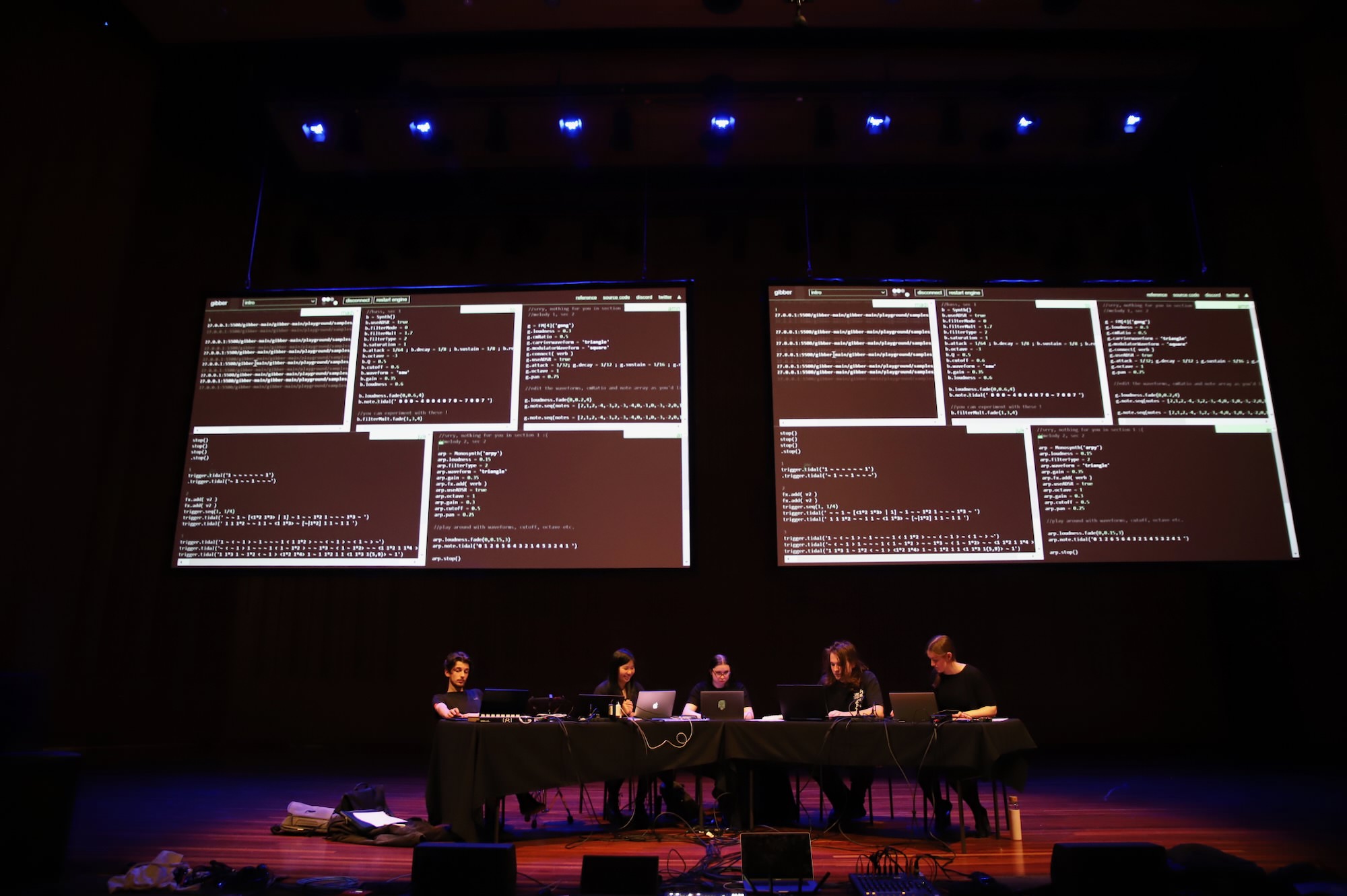 Five students playing laptops on desks in a large concert hall. Their code is projected on screens behind them.